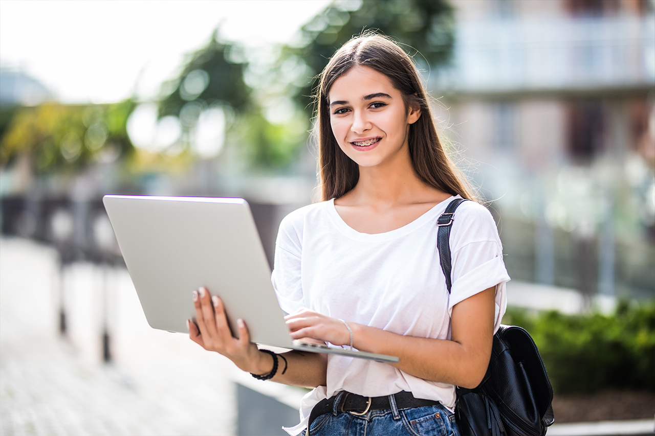Young woman with long hair and braces holding a laptop outdoors, wearing a white shirt and jeans.