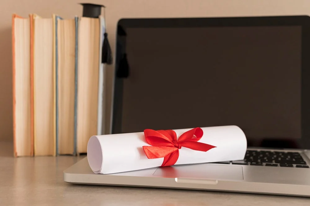 A rolled diploma tied with a red ribbon placed on a laptop next to stacked books and a graduation cap.