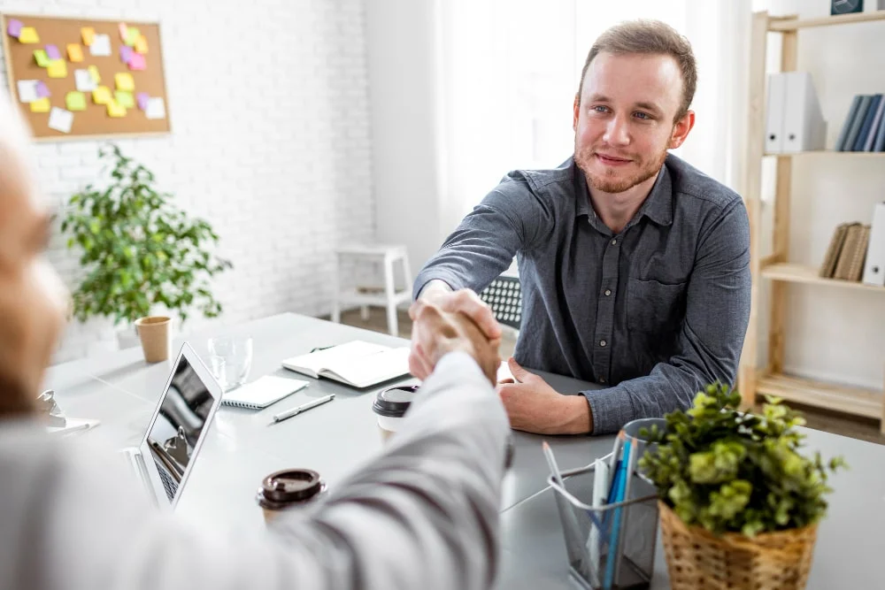 Two individuals shaking hands in a modern office setting during a job interview or meeting.