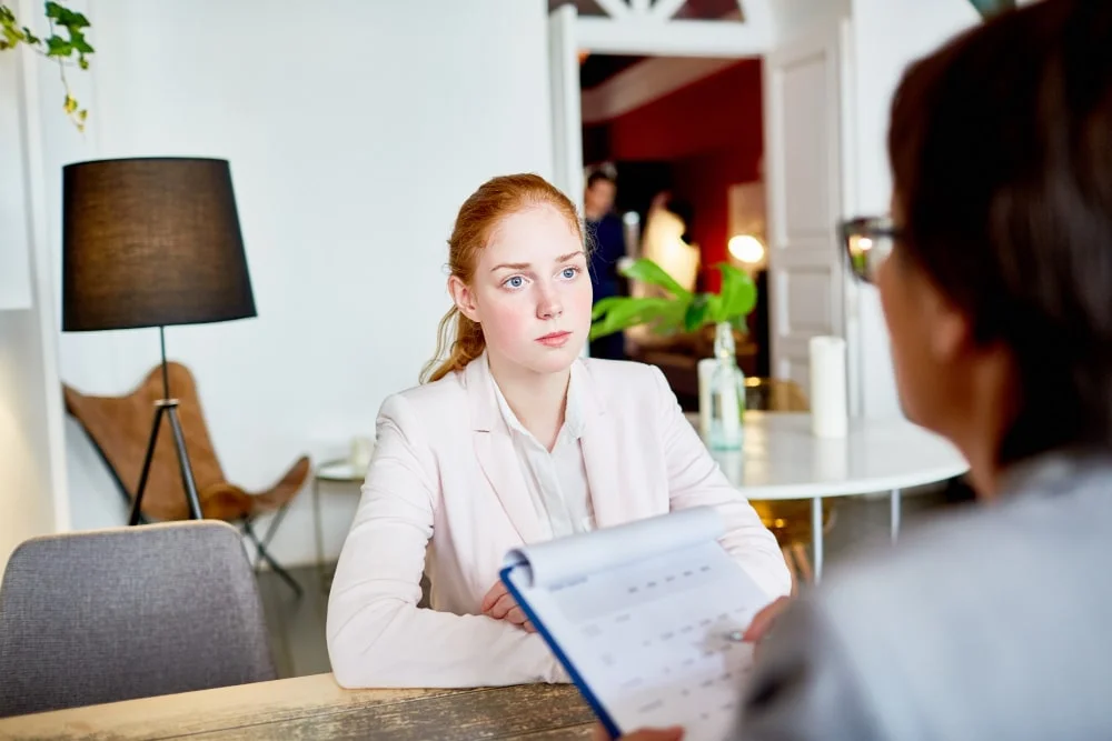 A young woman in a light-colored suit sitting across from a person holding a clipboard during a mock interview.