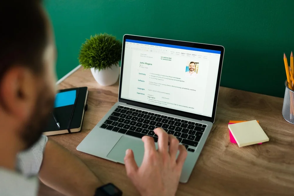 Individual reviewing a resume on a laptop screen with a plant and stationery on a desk.