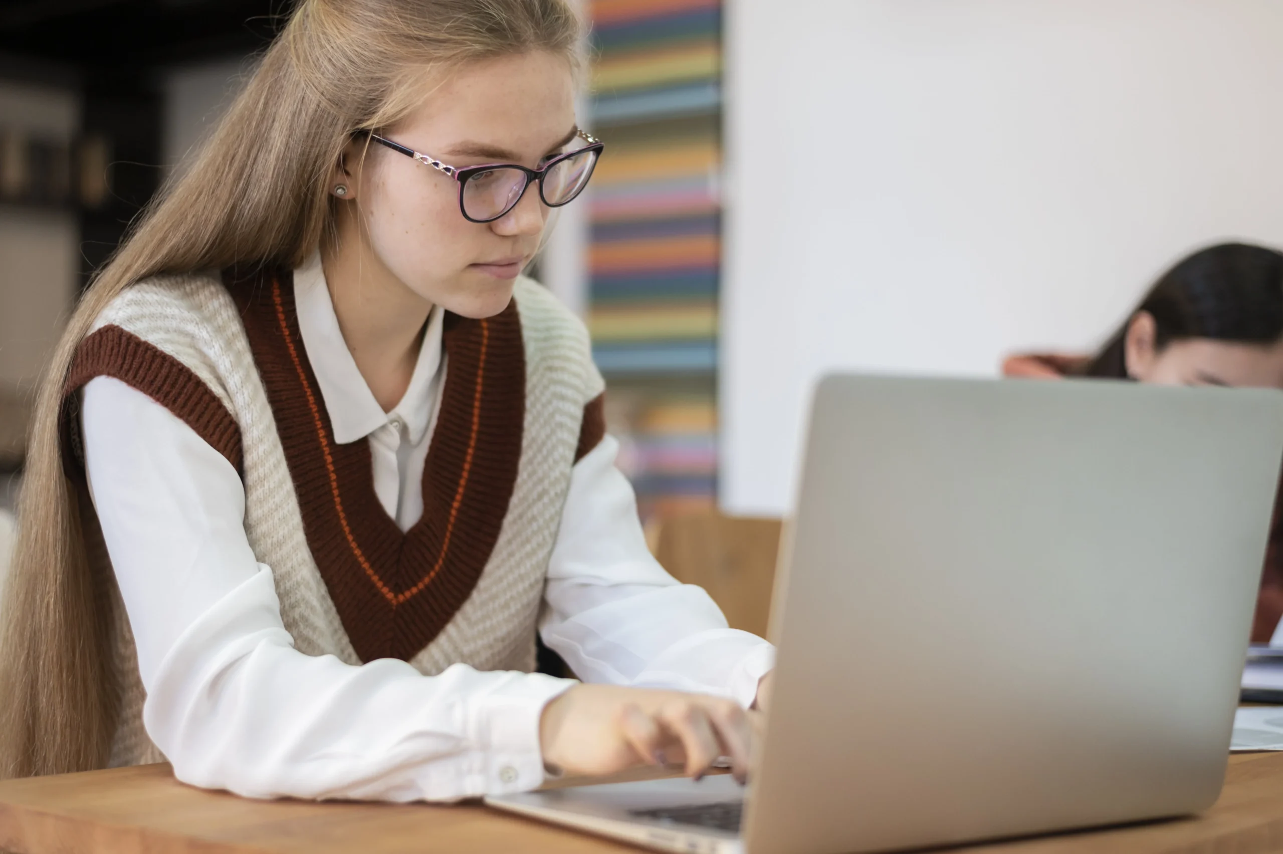 Young woman with long hair and glasses typing on a laptop while wearing a sweater vest in a study environment.